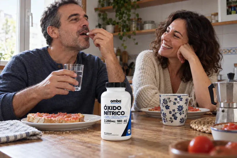 Pareja desayunando; él toma sus cápsulas con agua mientras ella bromea, el frasco de OMBA en la mesa